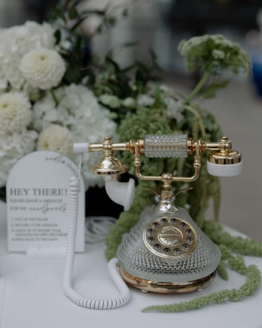 Decorative vintage-style phone with gold accents on a white surface with greenery and a card in the background.